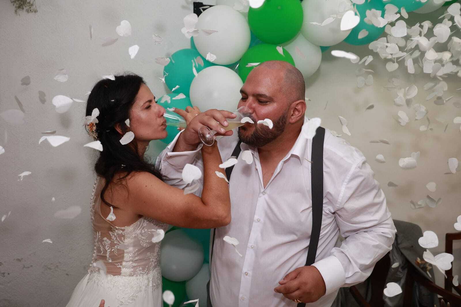Casal de noivos a brindar com champanhe rodeados de pétalas brancas a cair, com arco de balões verde ao fundo