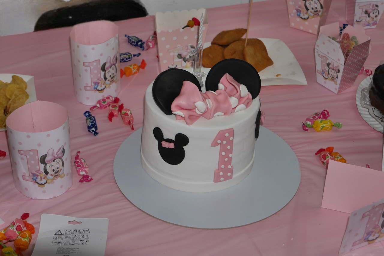 Bolo de primeiro aniversário decorado com tema Minnie Mouse sobre mesa rosa com doces e material gráfico personalizado